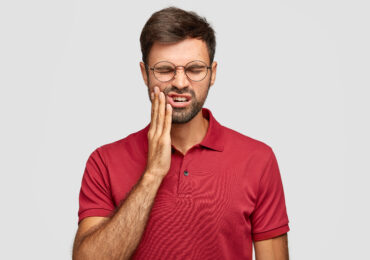 Photo of Caucasian man suffers painful toothache, has rotten tooth, needs to visit dentist, keeps eyes closed from pain, dressed in casual red t shirt, isolated over white background, touches cheek