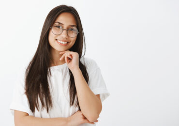 Waist-up shot of beautiful dreamy girlfriend in stylish eyewaer, holding hand near chin with hand crossed on chest, smiling broadly and tilting head while listening carefully with joy over gray wall.