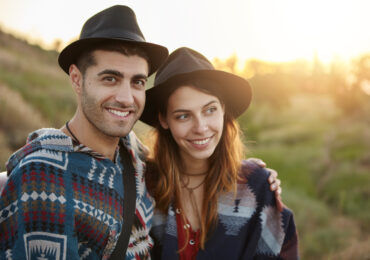 Couple of beautiful girlfriend and boyfriend in black hats sitting on grass at meadow smiling and embracing admiring nature and fresh air talking with each other. Romantic atmosphere, relaxation